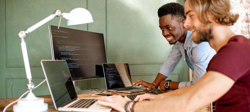 Two young programmers working with program code sitting at the workplace with three monitors in the office on the green wall background